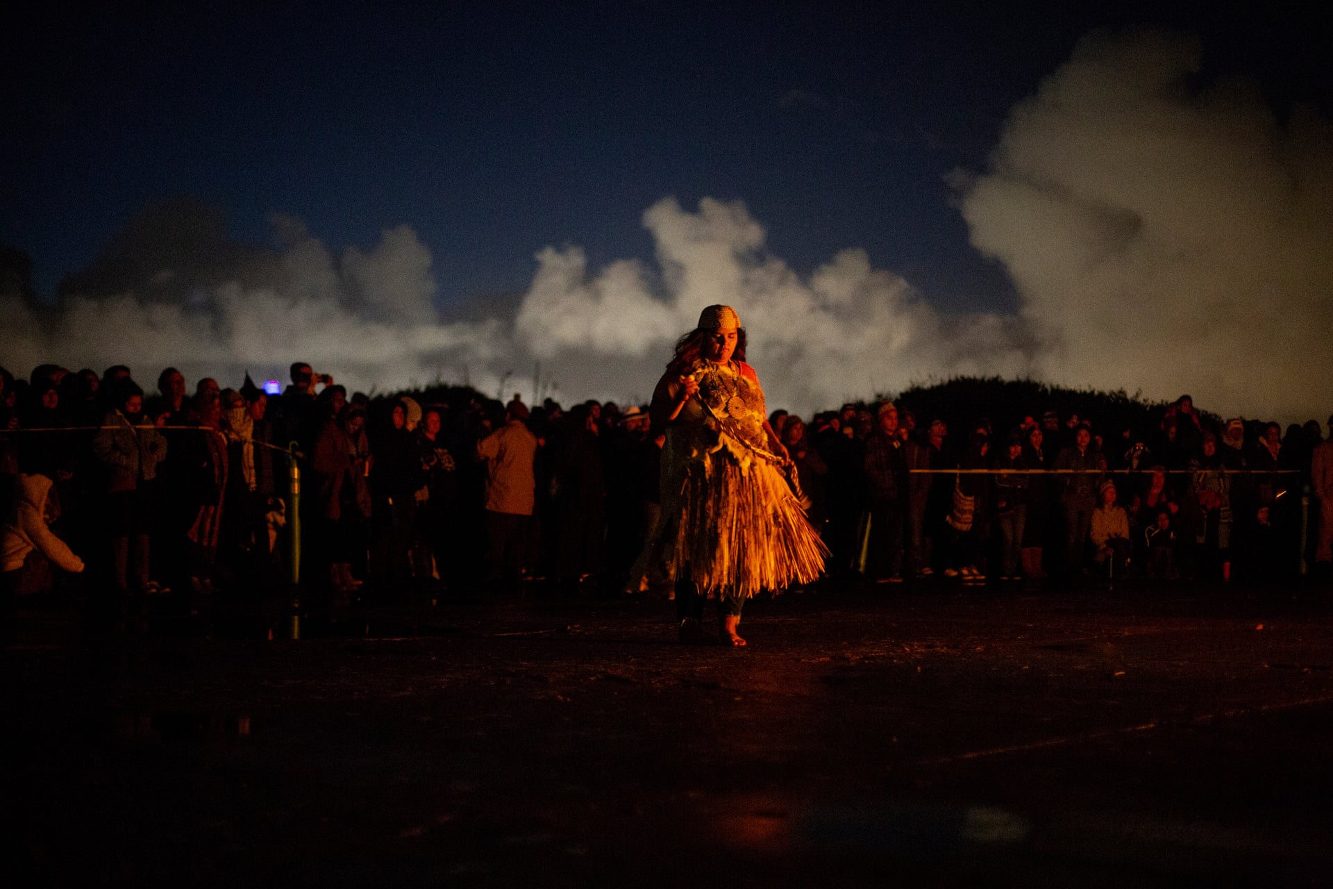Color photograph showing a large crowd outside. A female dancer in Native cultural clothing dances at the center of the image.